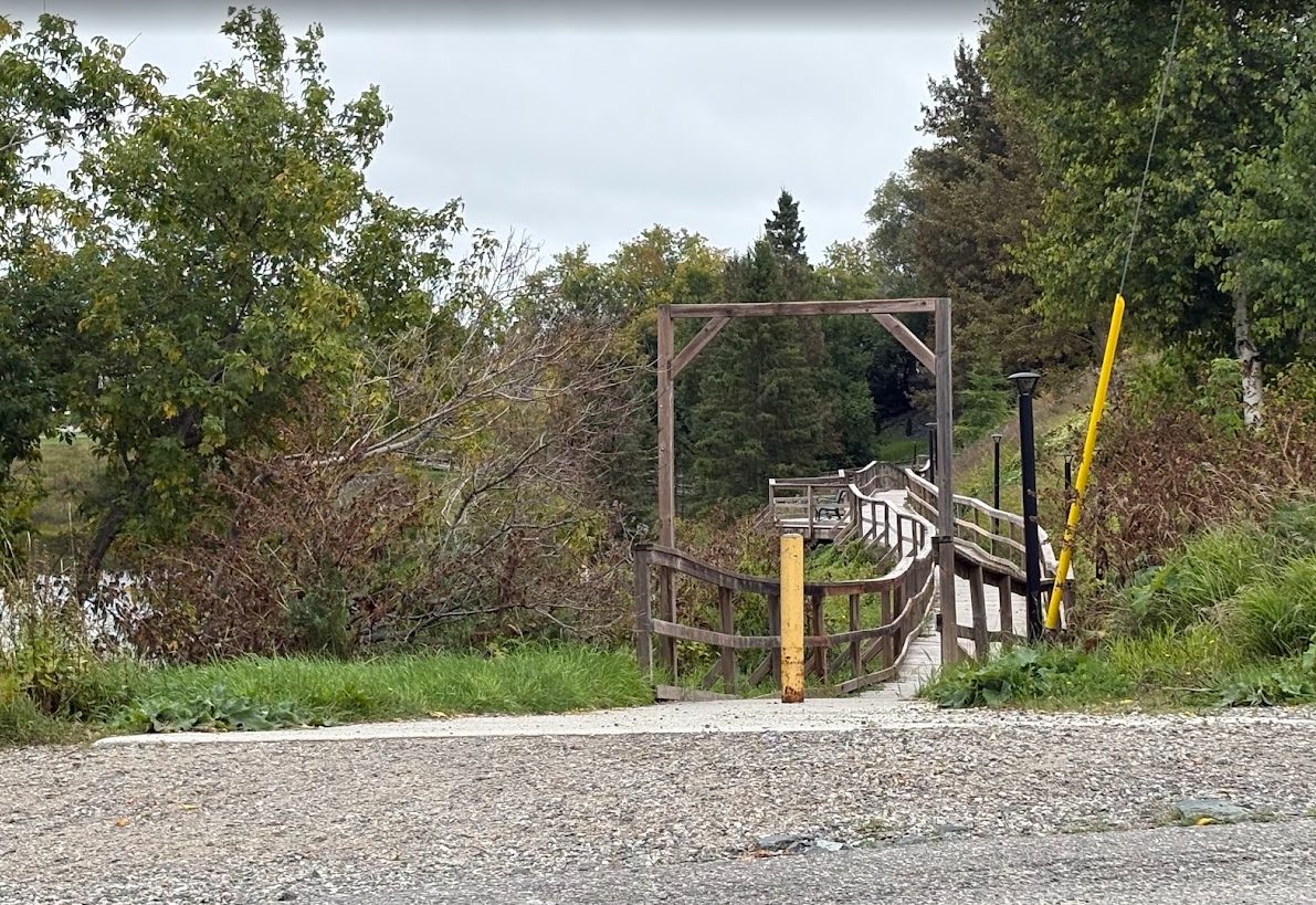 Brush and weeds crowding the wooden archway at the entrance to the Lake Commando boardwalk in Cochrane